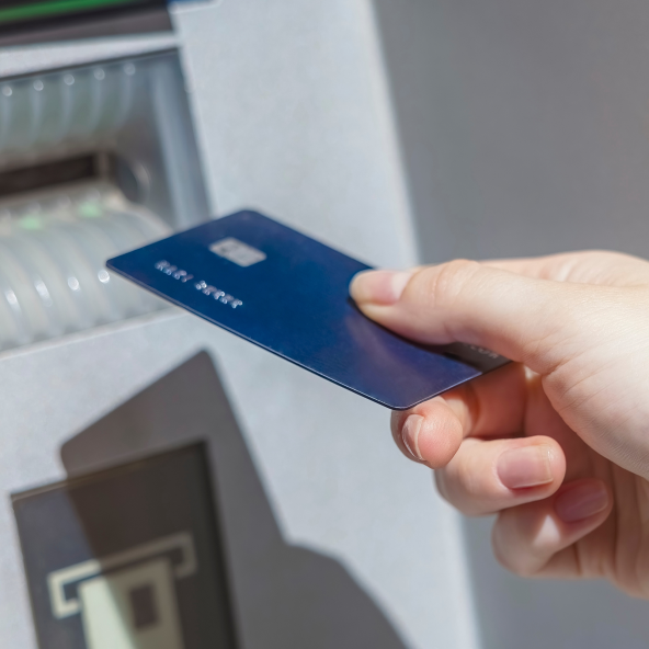 Dłoń trzyma w ręku niebieską kartę płatniczą i zbliża ją do bankomatu
